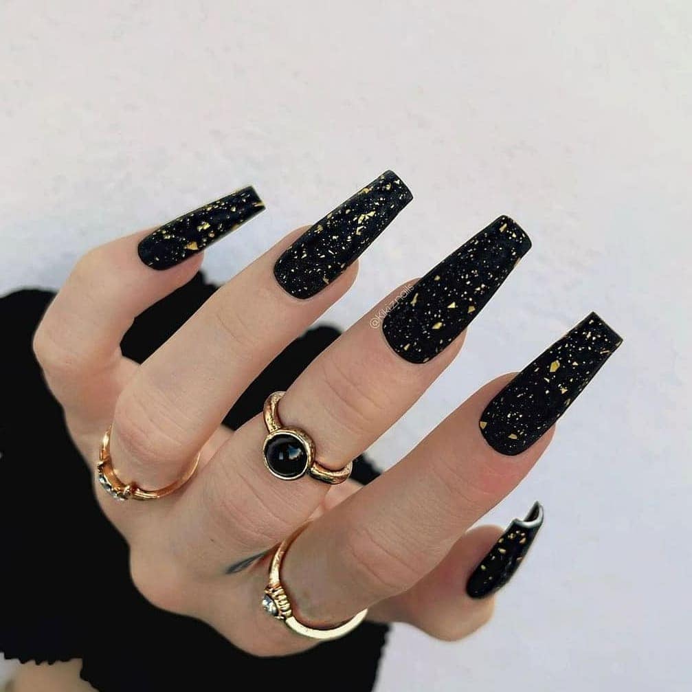 hand wearing gold rings and long coffin-shaped black nails with gold glitter flakes.