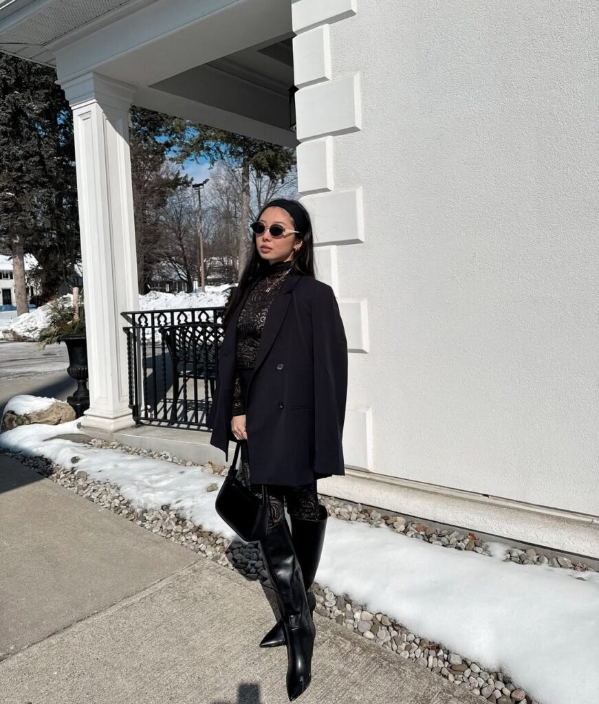 woman in a black lace jumpsuit, oversized blazer, and tall black leather boots in snow.