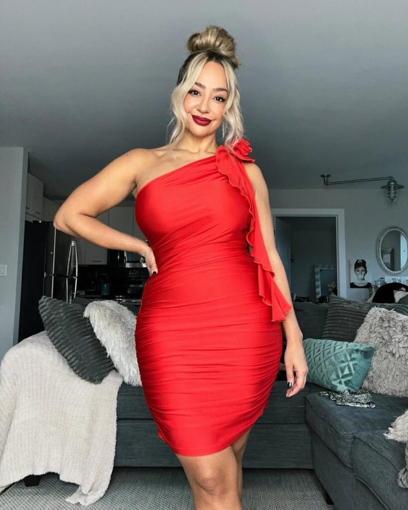 woman in a red one shoulder ruffled mini dress posing in a living room.