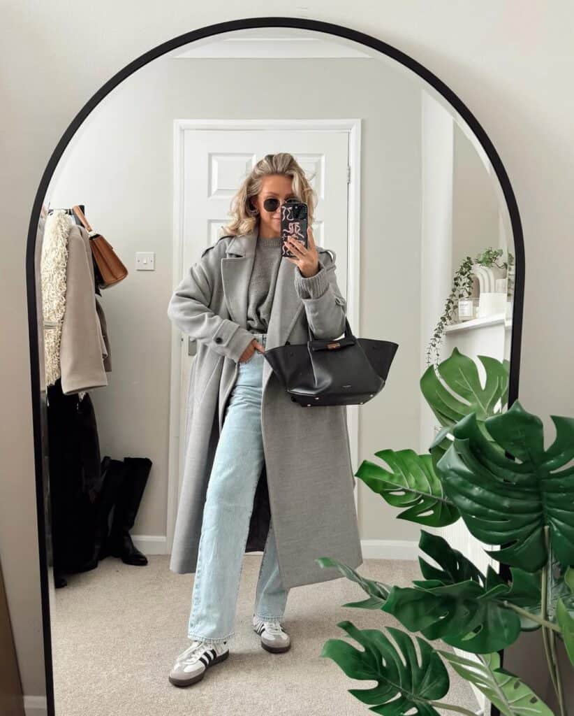 blonde woman wearing a long grey coat, charcoal sweater, and light wash jeans taking a mirror selfie for spring outfits inspo