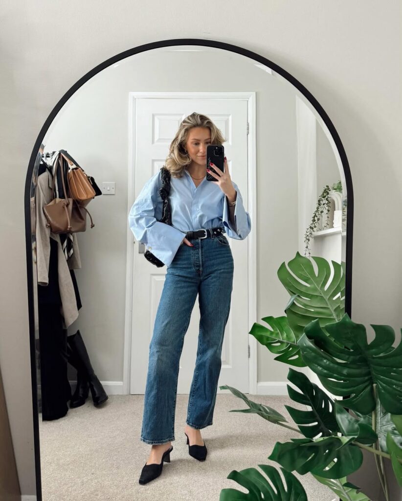 blonde woman wearing a light blue oversized button-down shirt and straight-leg jeans taking a mirror selfie