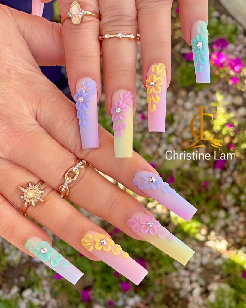 Long acrylic nails with a gradient design featuring pastel colors and floral embellishments, adorned with small rhinestones.