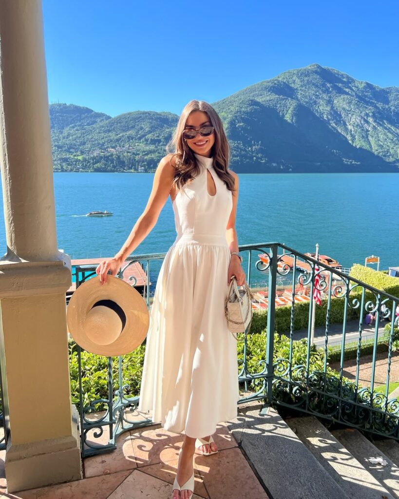 woman in a sleeveless white midi dress stands on a balcony overlooking a lake