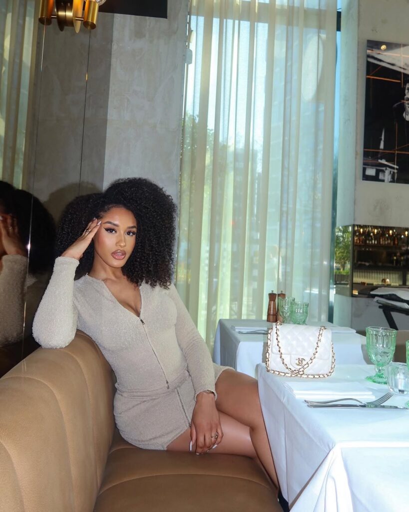 Woman with curly hair seated on a couch in a restaurant, wearing a fitted beige dress, with a white handbag beside her.