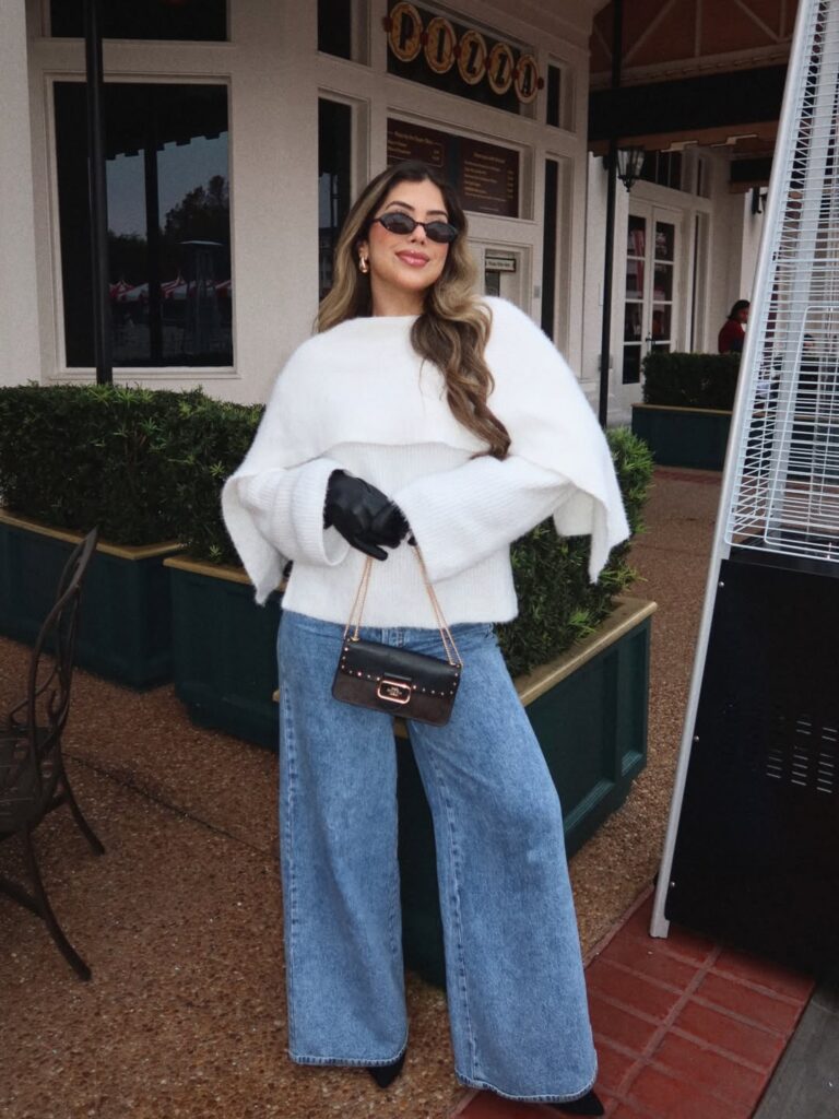 Woman wearing a white sweater and wide-leg denim jeans, holding a small handbag, with sunglasses and gloves.