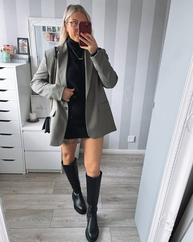 Person wearing a gray blazer, black top, and knee-high black boots, taking a mirror selfie in a well-lit room.
