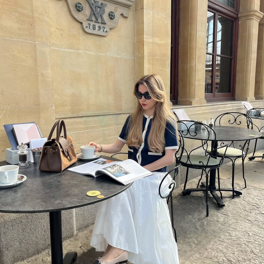 woman in a navy short-sleeved cardigan and white midi skirt sitting at an outdoor cafe
