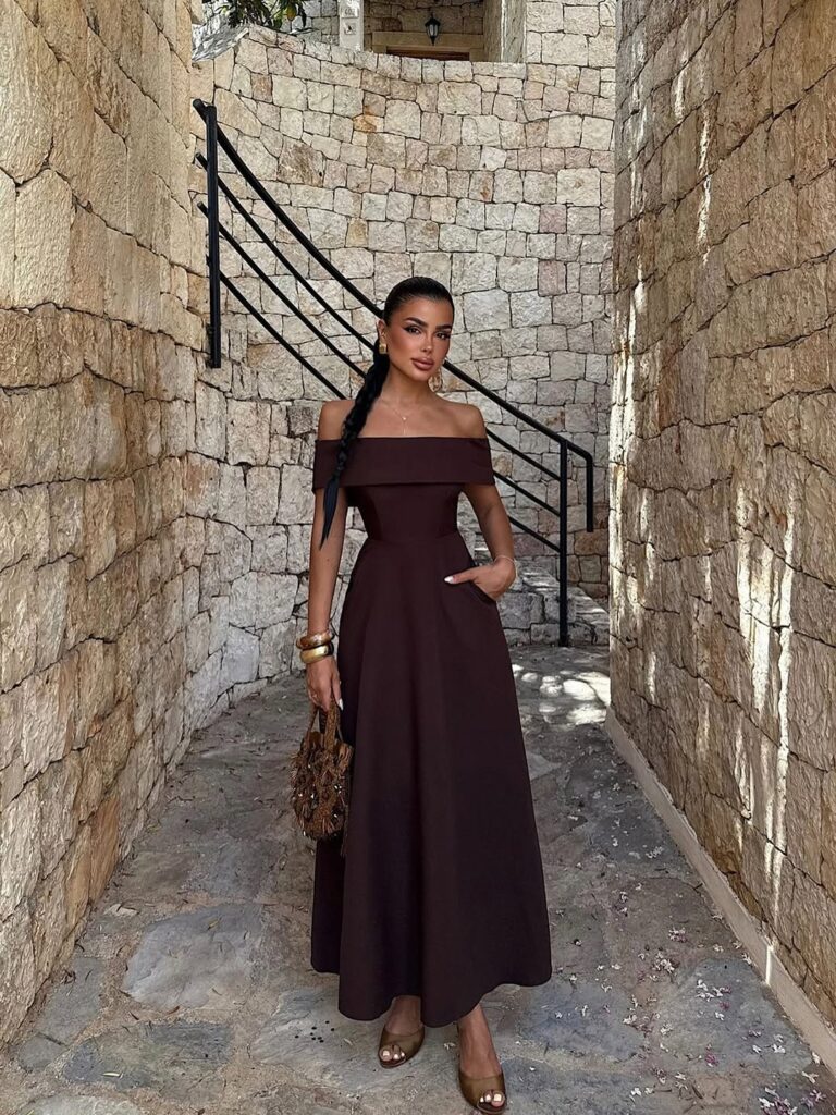 woman in brown off-the-shoulder maxi dress holding a beaded handbag between stone walls