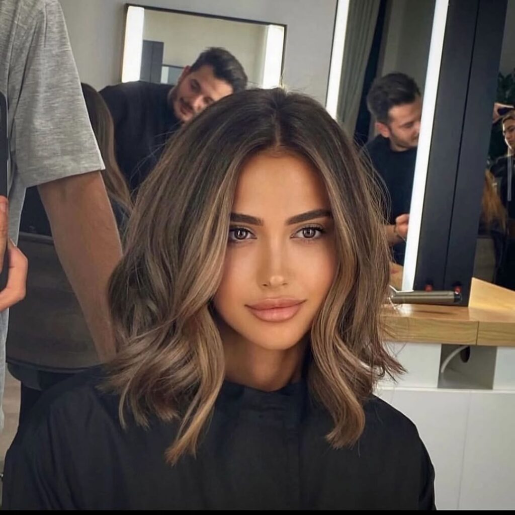 Woman with medium-length, layered hair styled in soft waves, seated in a salon chair with stylists in the background.
