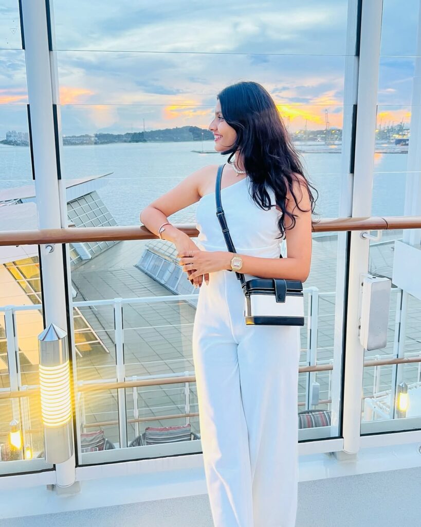 woman in a white one shoulder jumpsuit leaning on a cruise ship railing at sunset
