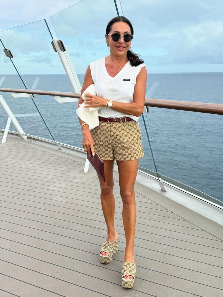 smiling woman in white v neck shirt and patterned shorts walks on cruise ship deck
