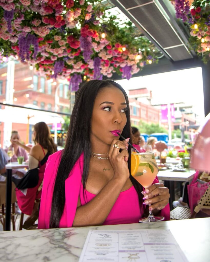 Woman in a pink outfit sipping a colorful cocktail with fruit garnishes in a vibrant restaurant setting.