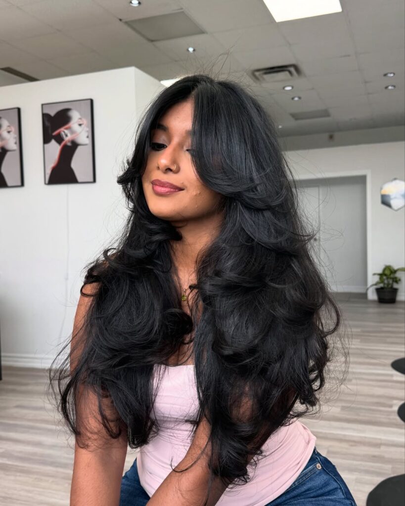 Woman with long, voluminous black hair styled in loose waves, wearing a light pink top and sitting in a salon environment.