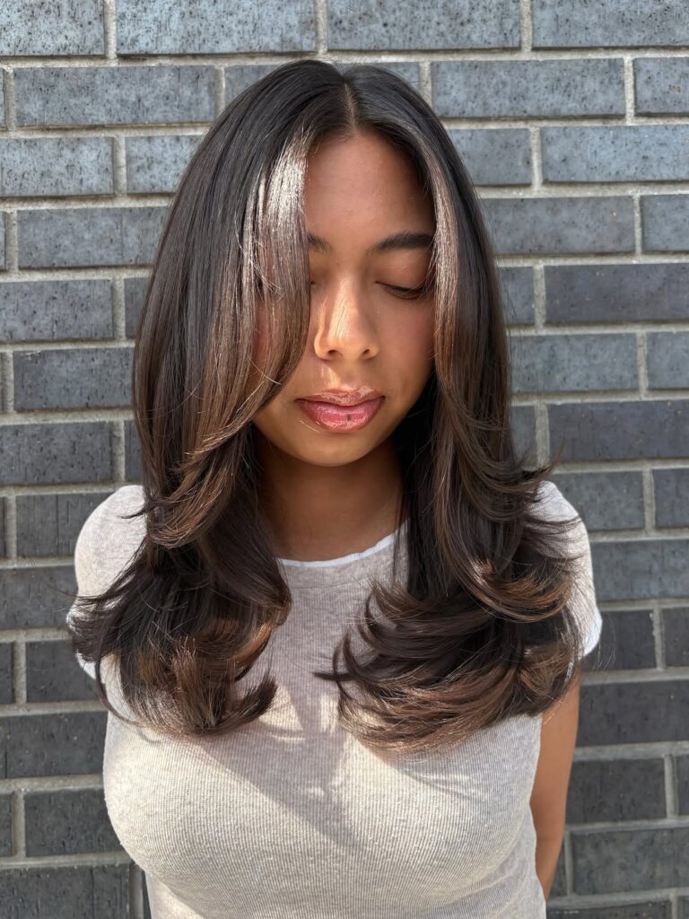 Woman with long, layered hair styled with soft waves, standing against a gray brick wall.