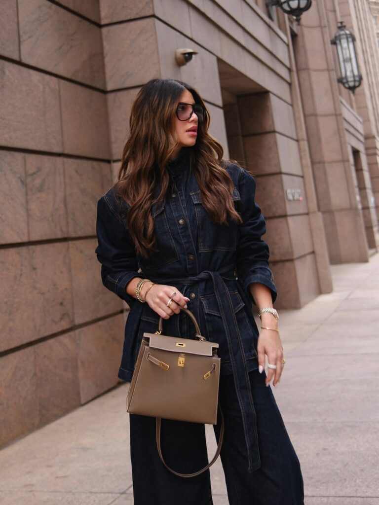 woman in dark denim belted jumpsuit and sunglasses holding a tan leather handbag outdoors