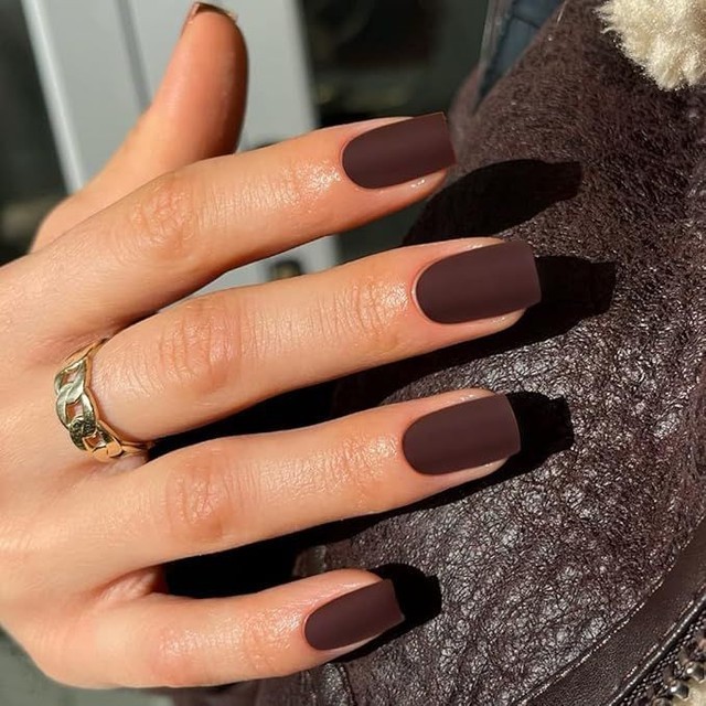 Hand with long, matte brown nails resting on a textured surface, adorned with a gold ring on the finger.