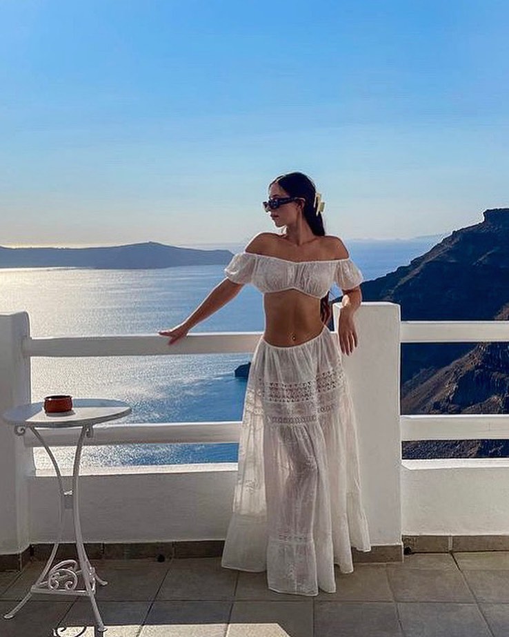 woman in a white two piece linen outfit stands on a balcony overlooking the sea