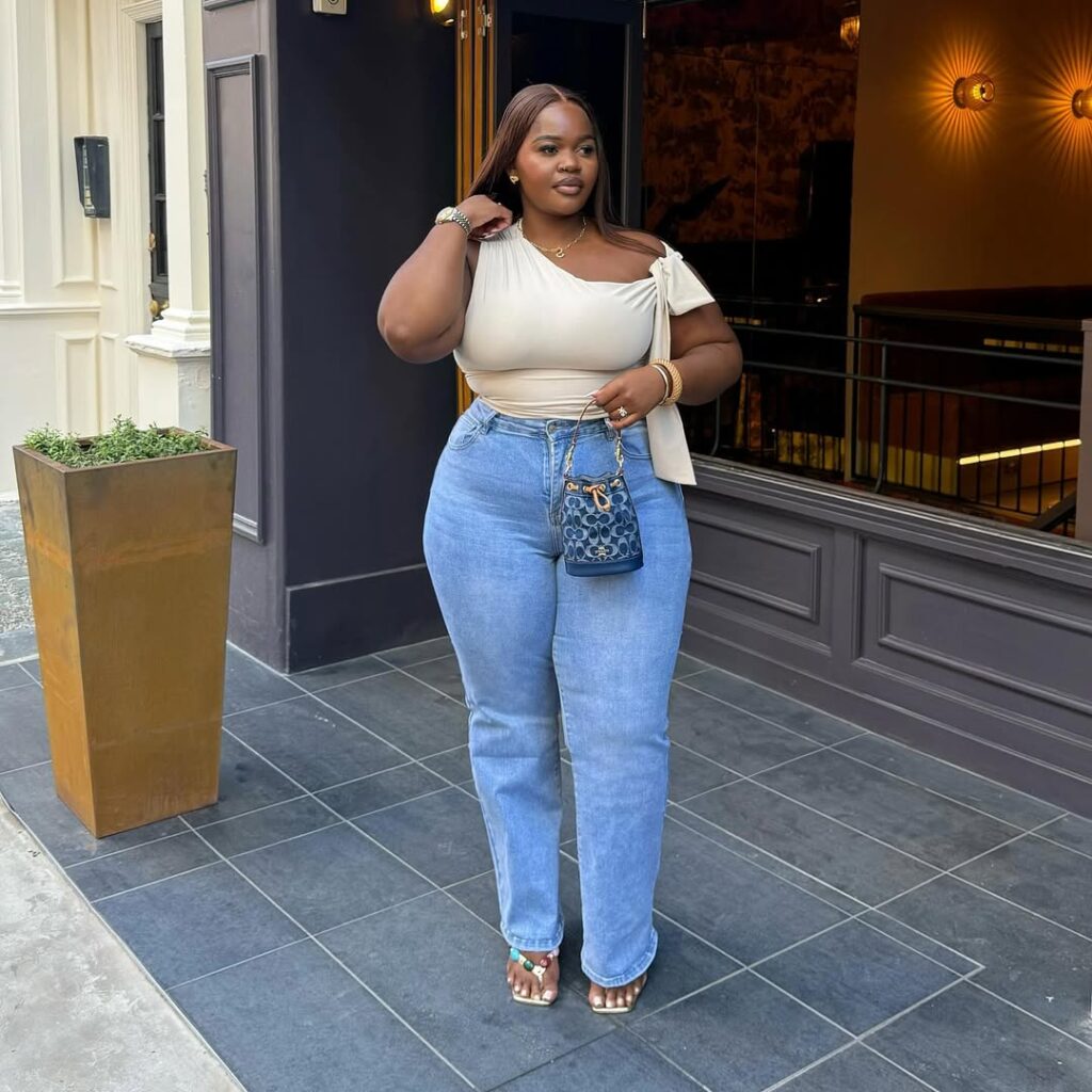 Woman wearing a light-colored off-shoulder top and blue jeans, standing outside a building with a stylish entrance.