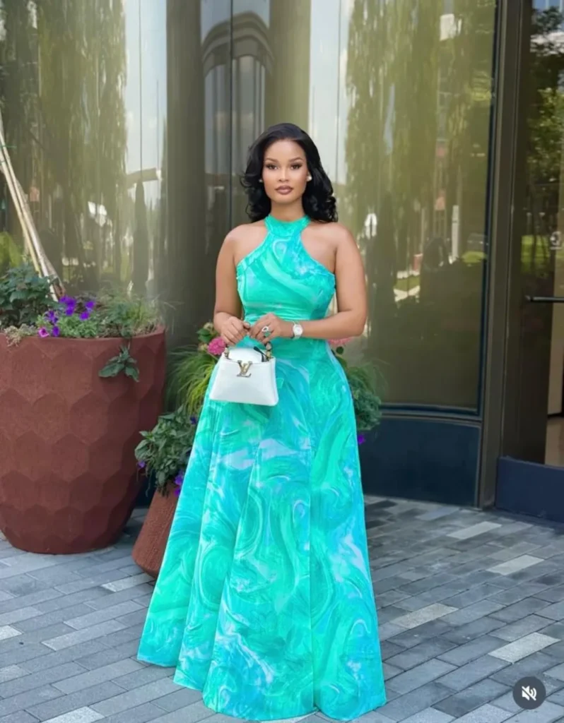 Woman wearing a green marble-patterned dress with a halter neck design, holding a white handbag, standing outside a modern building.
