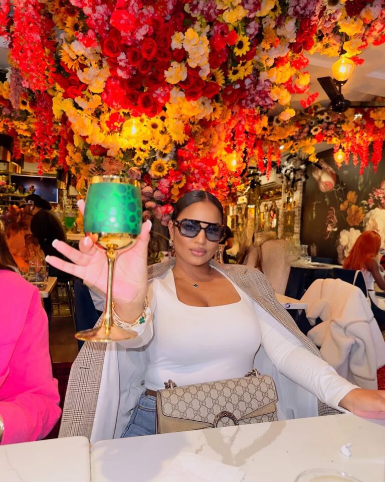 Woman holding a green decorative glass in a vibrant setting adorned with colorful flowers and floral arrangements.