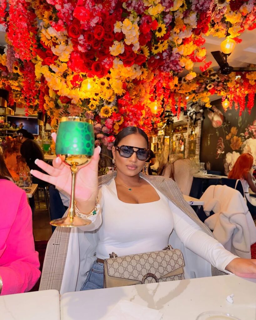 Woman holding a green decorative glass in a vibrant setting adorned with colorful flowers and floral arrangements.