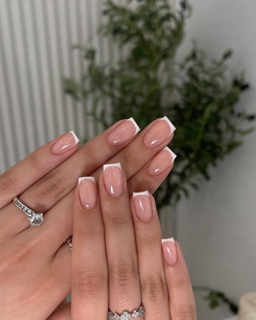Hands with manicured nails featuring a nude base and white French tips, adorned with rings on the fingers.