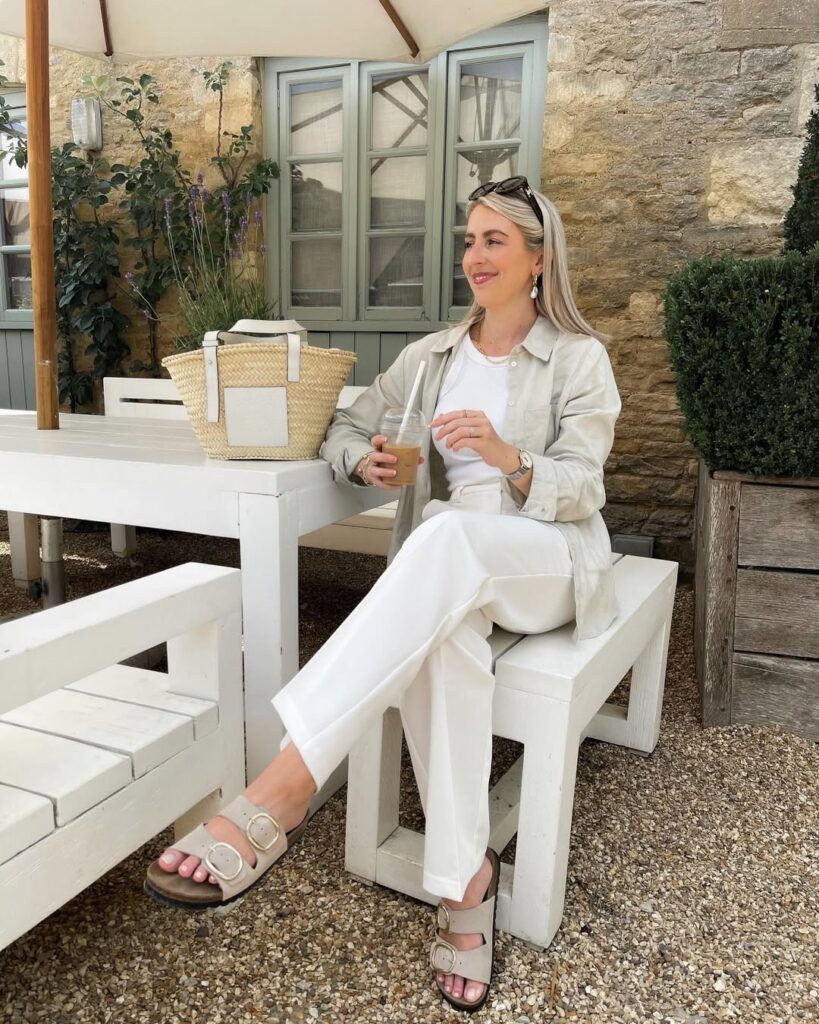blonde woman sitting at white outdoor table wearing linen shirt and trousers holding iced coffee