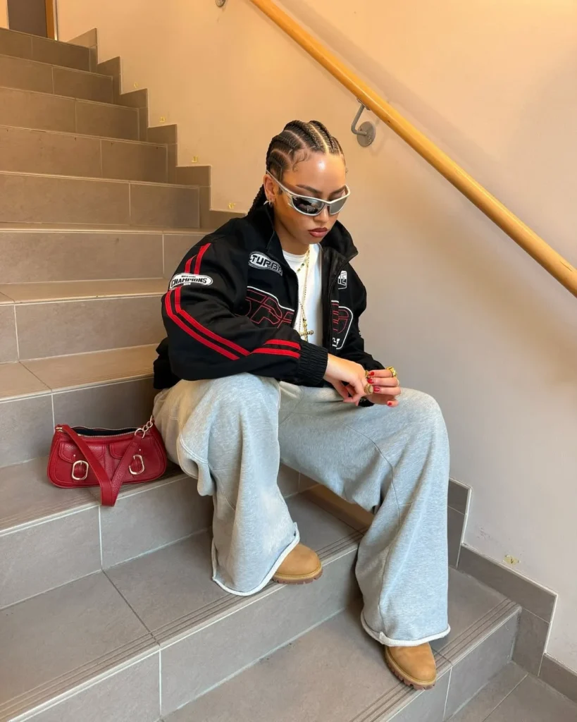 woman in a racing jacket and oversized grey sweatpants sitting on indoor stairs