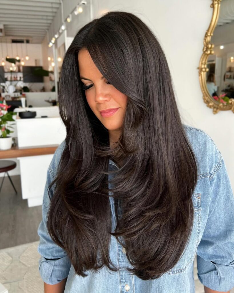 Woman with long, smooth dark hair styled in soft waves, wearing a denim shirt in a salon setting.