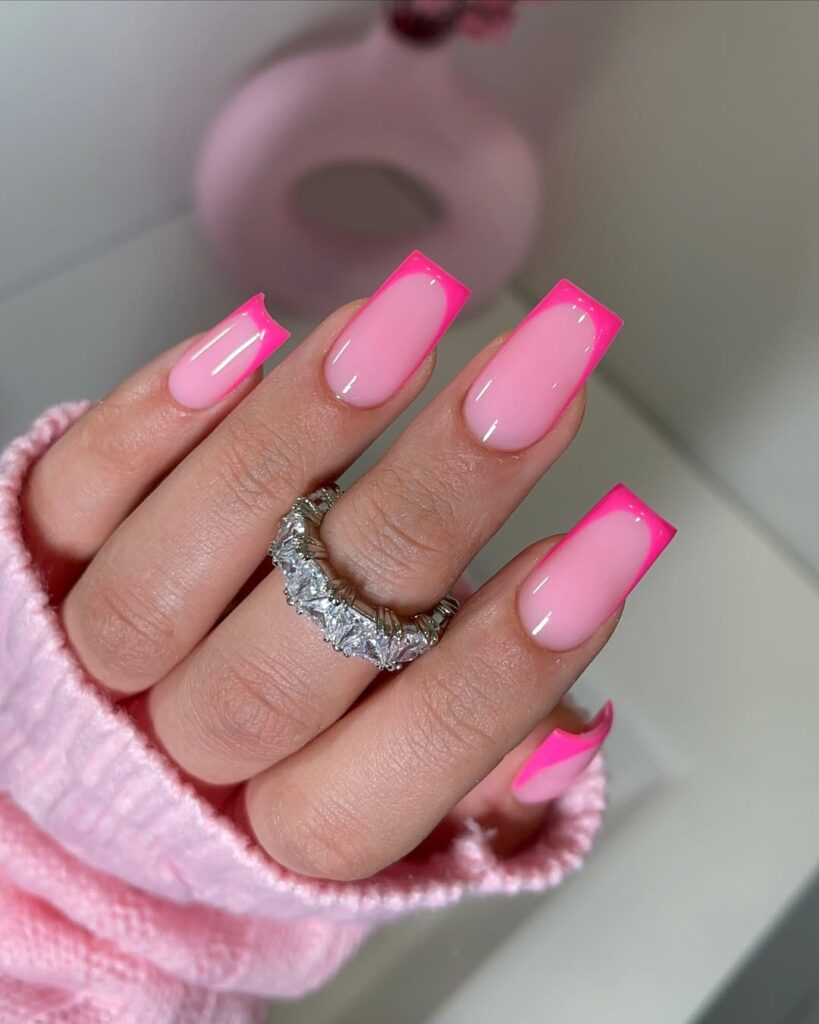 Hand with long pink nails featuring bright pink tips, adorned with a silver ring, against a soft background.