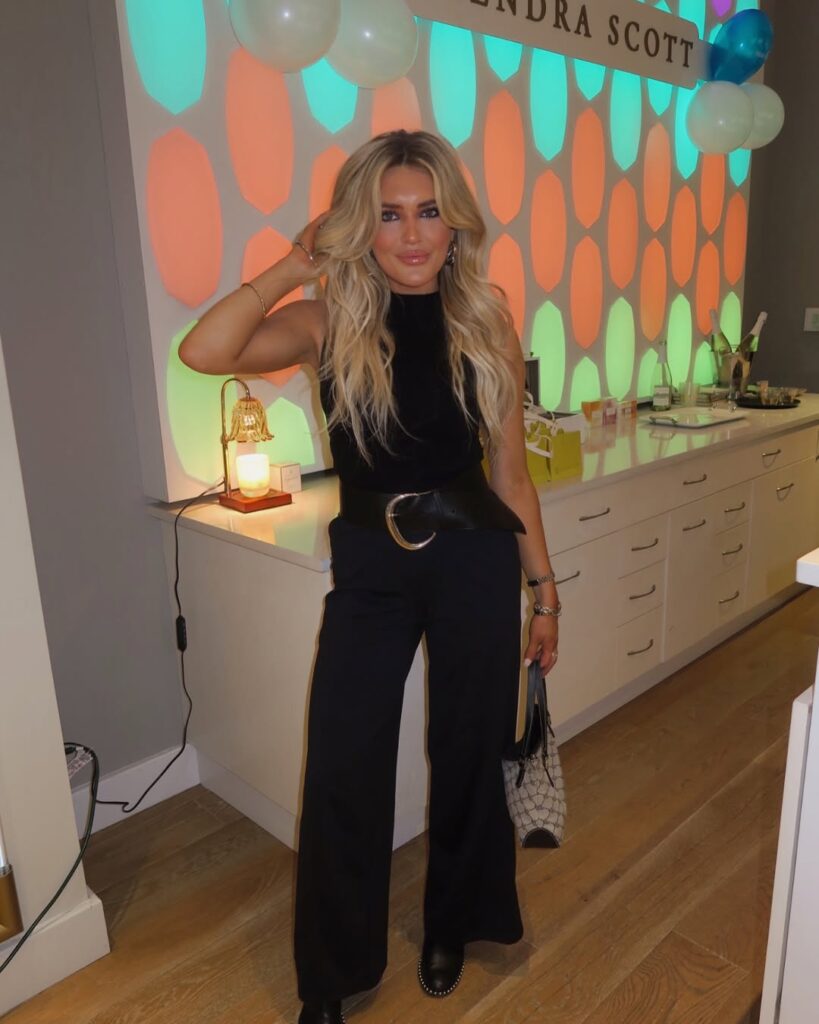 Woman with long blonde hair wearing a black outfit and holding a handbag, standing in a brightly lit room with colorful wall decor.
