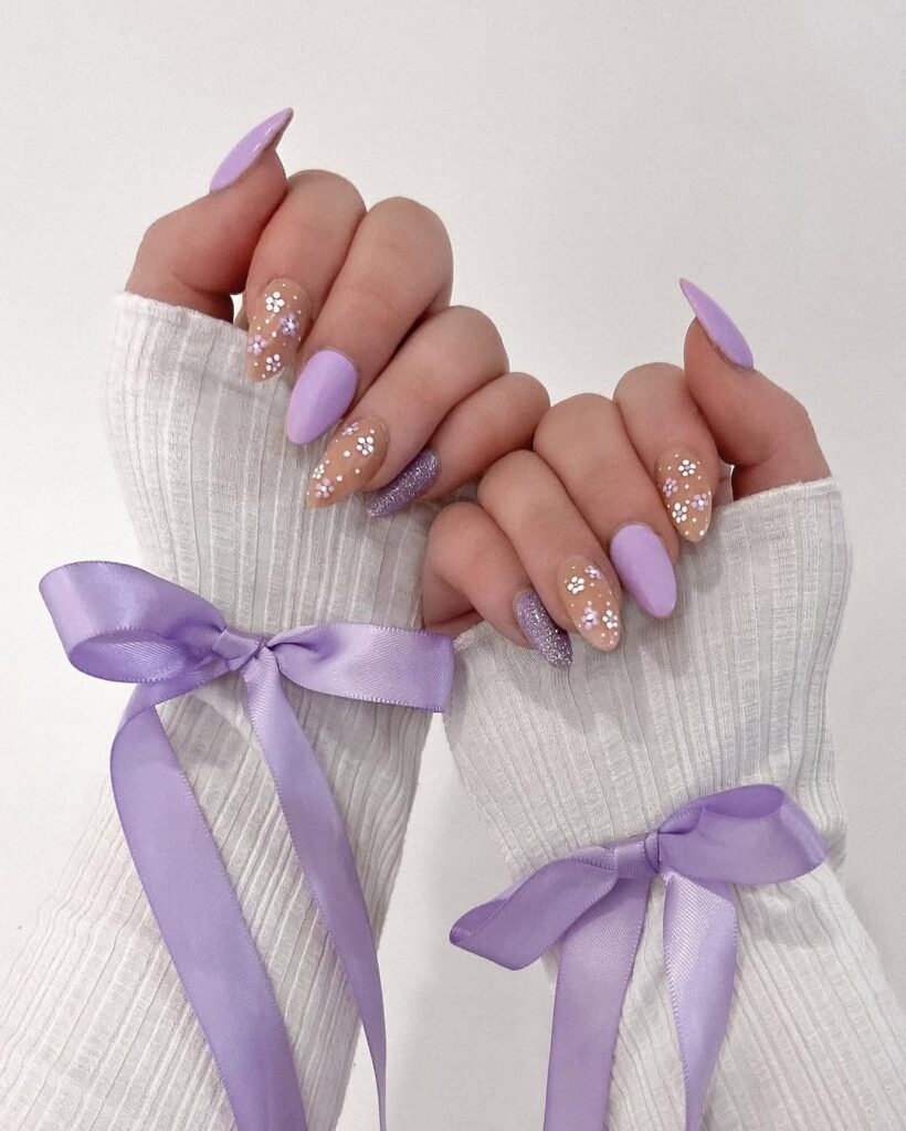 Hands with long lavender nails adorned with glitter and floral designs, wearing white fingerless gloves with lavender ribbons.