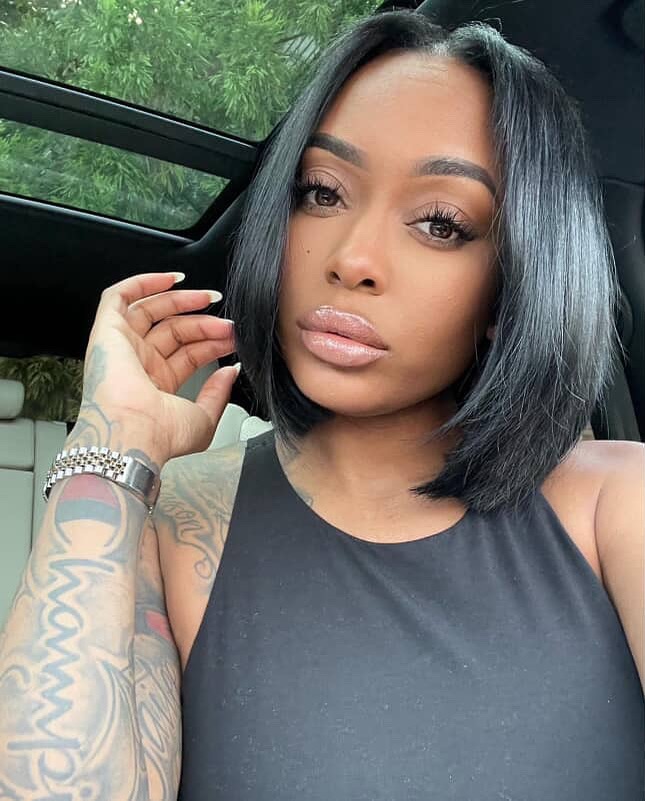 Woman with shoulder-length black hair and a black sleeveless top, showing tattoos on her arm, posing inside a car.