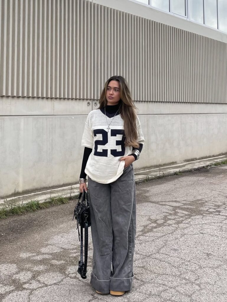 woman in a numbered jersey and wide grey trousers standing against a ribbed wall