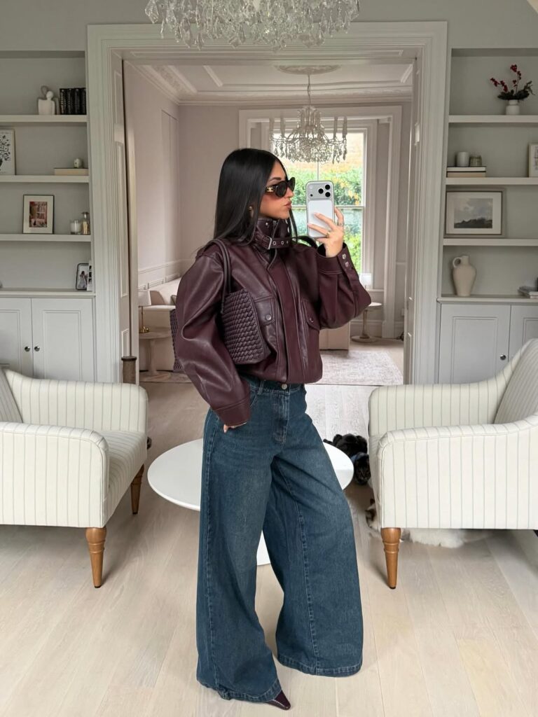 Woman wearing a burgundy leather jacket and wide-leg denim jeans, posing in a modern interior with a stylish handbag.
