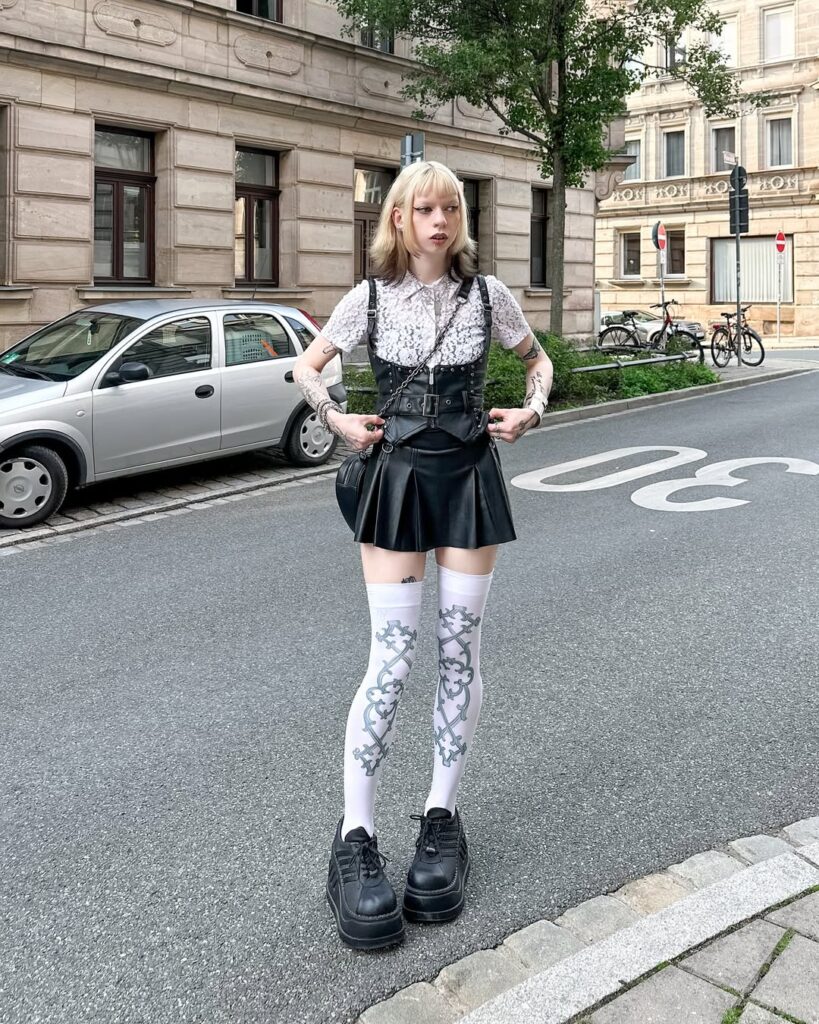 blonde woman wearing a white lace shirt, black leather corset, and pleated mini skirt