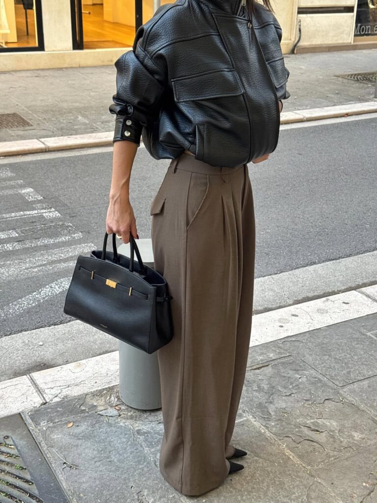 Woman wearing a black leather jacket and brown wide-leg trousers, holding a black handbag with gold accents.