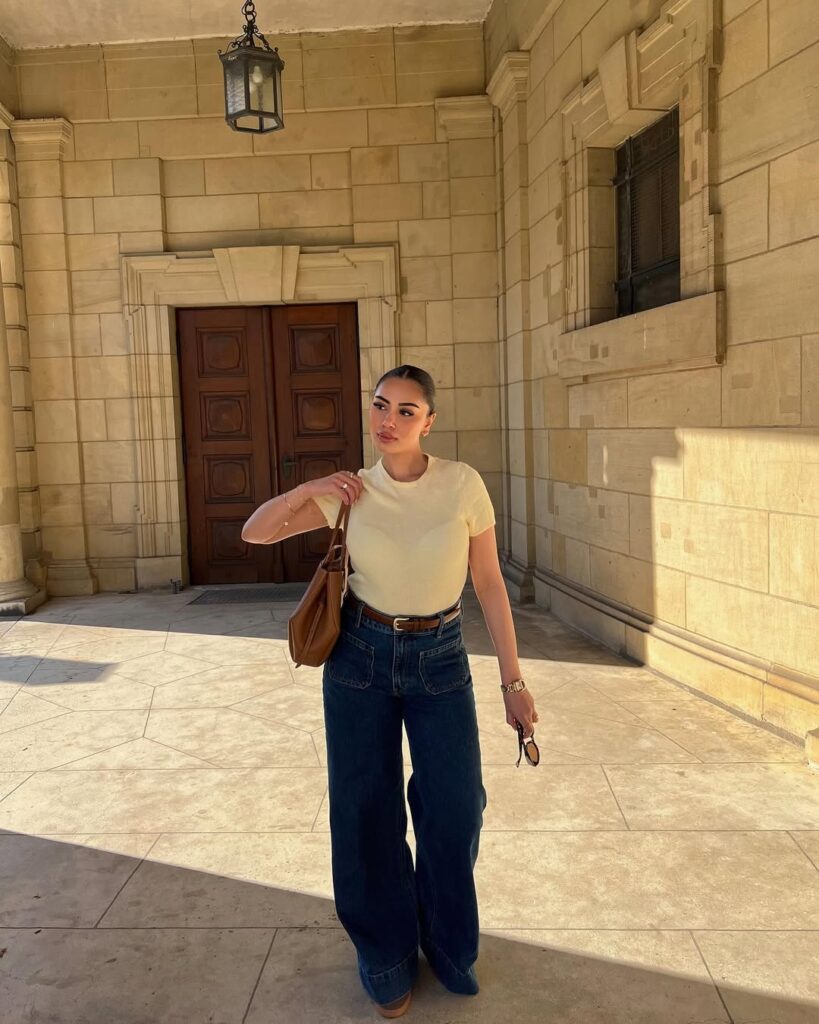 woman in a pale yellow t-shirt and dark wide-leg jeans posing in front of stone architecture