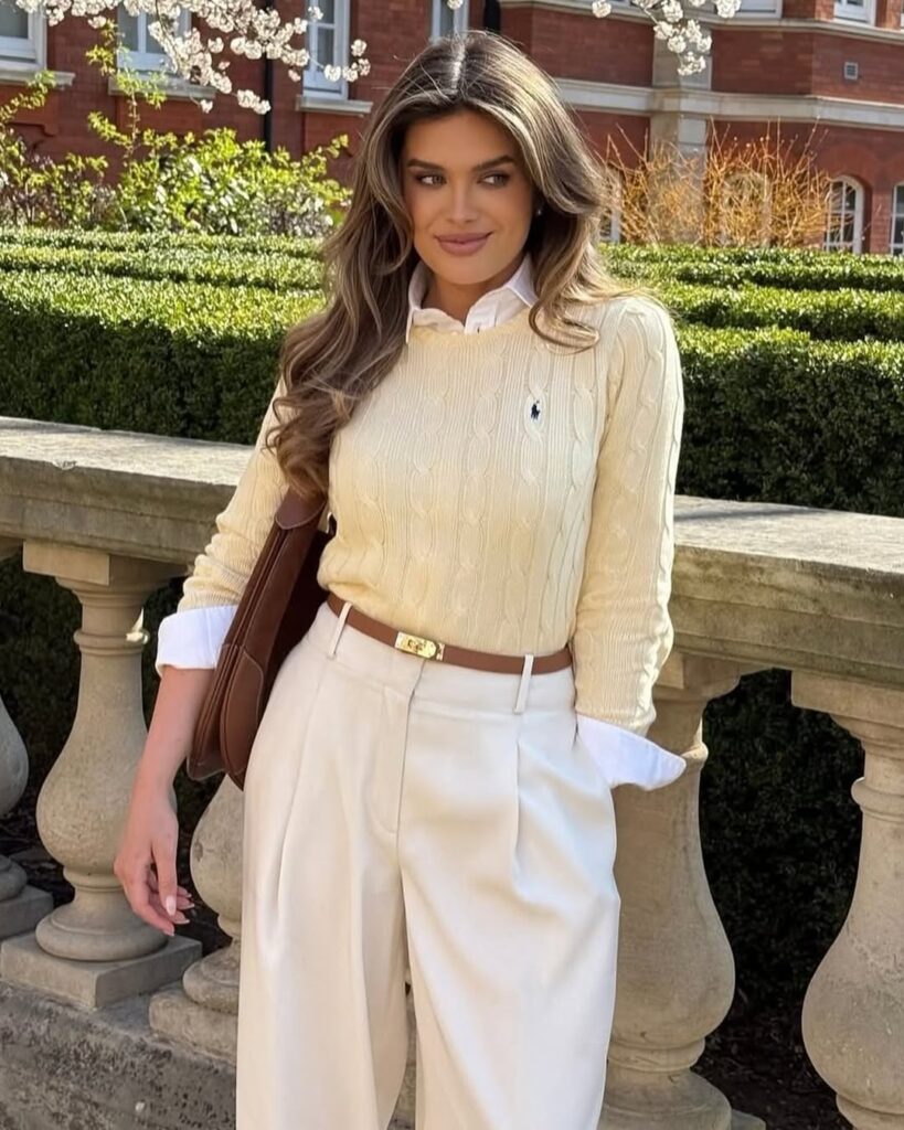 woman in a cream cable knit sweater and white trousers posing outdoors by a stone railing