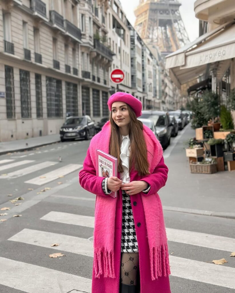 woman in pink coat, beret, and houndstooth skirt holding a book on a paris street.