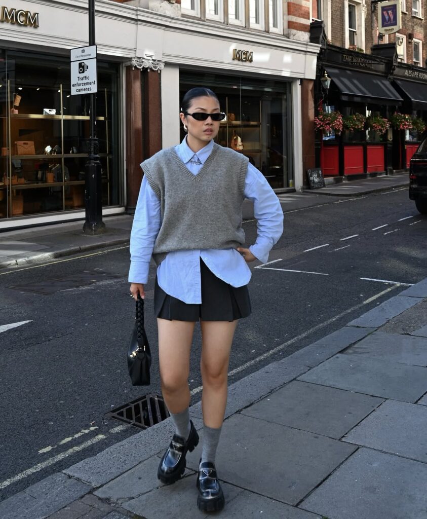 woman in a grey sweater vest and blue button-down shirt walking on a city sidewalk for spring outfits inspo