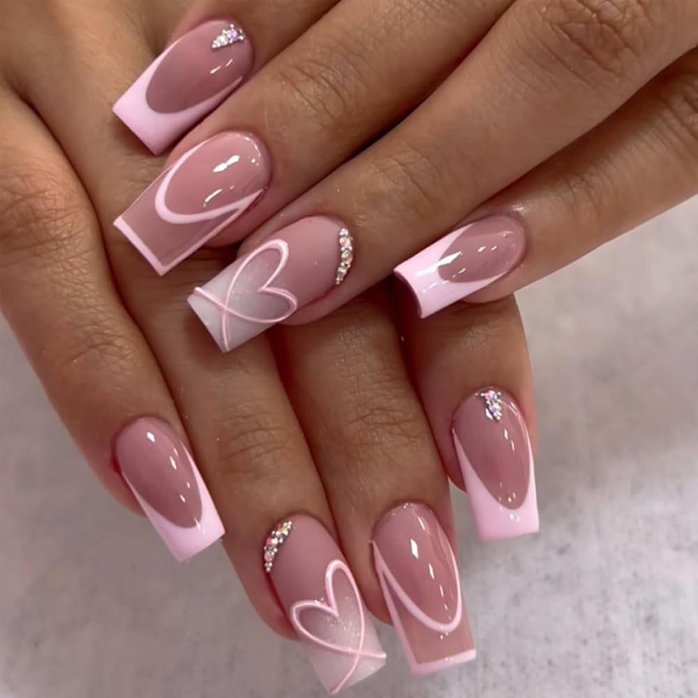 Nail art design featuring long square-shaped nails with a nude base, pink tips, and white heart patterns with glitter accents.