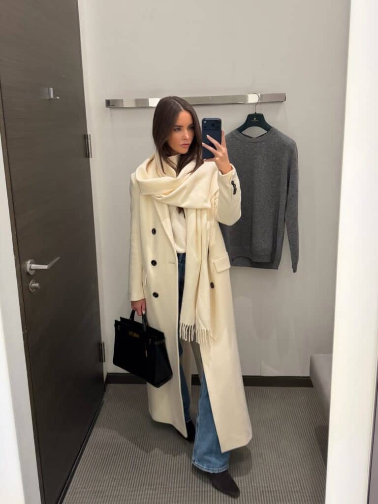 woman in long cream coat and matching scarf taking mirror selfie in dressing room