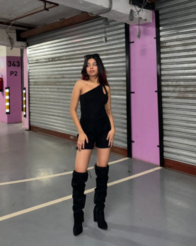 woman in black one-shoulder romper and knee-high boots posing in a purple parking garage