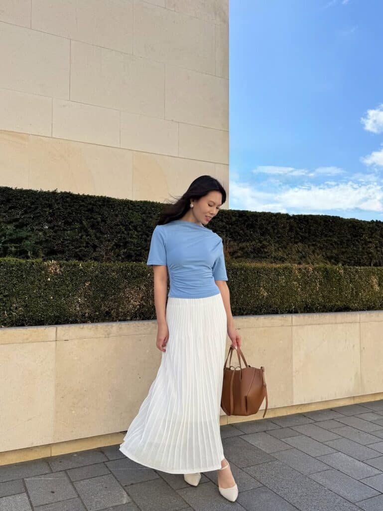 woman in light blue top and white pleated skirt holding a brown leather bag
