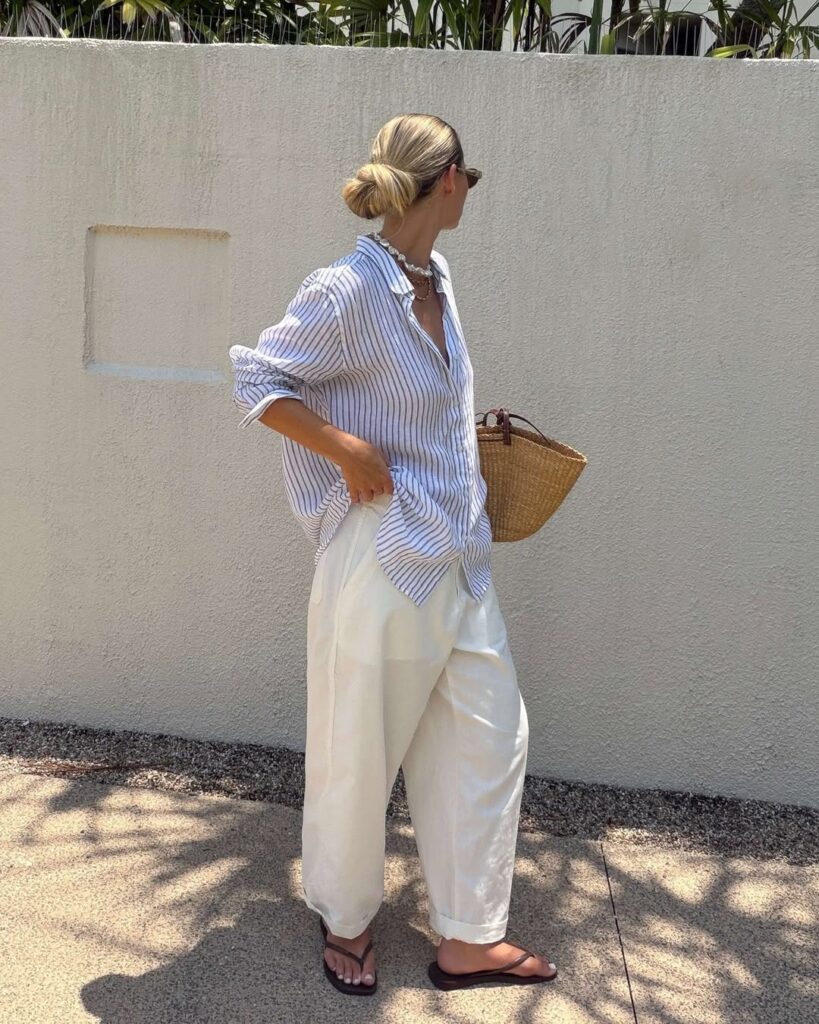 woman in blue striped linen shirt and white trousers carrying a woven straw bag