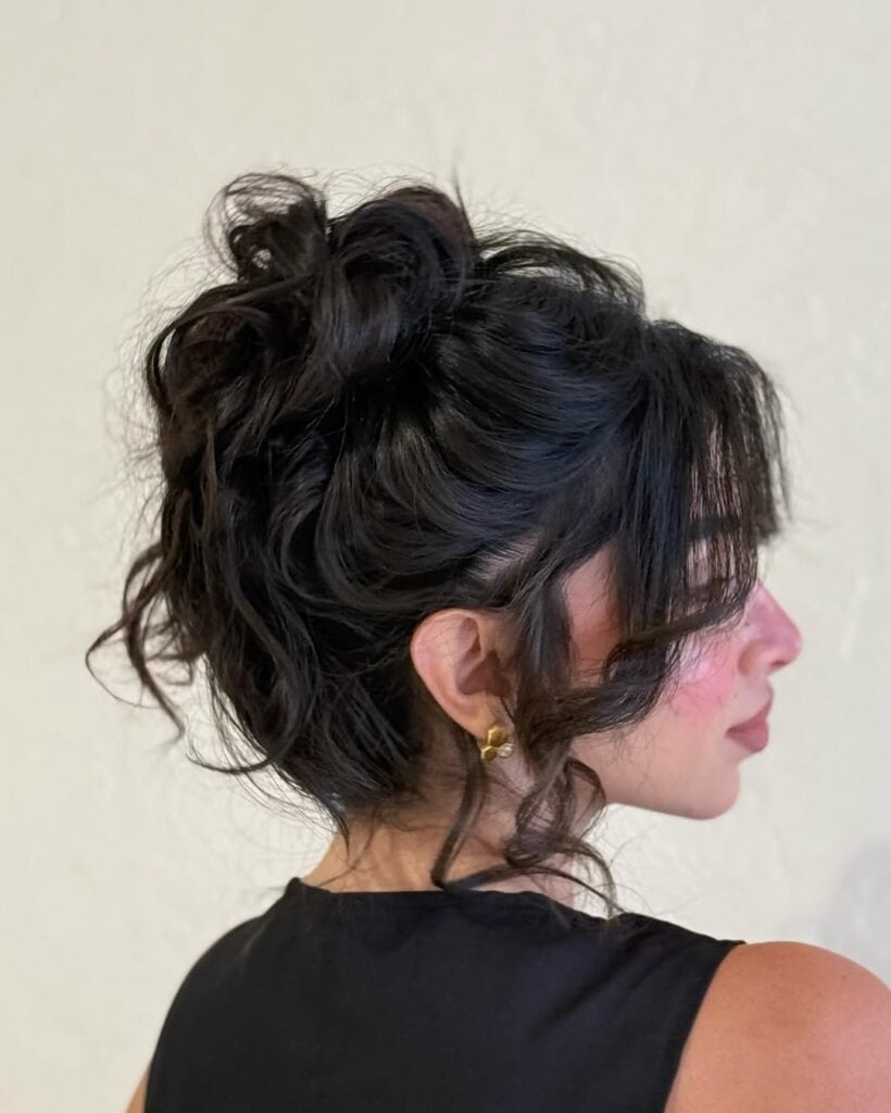 messy dark brunette updo with curly face-framing fringe and gold stud earrings