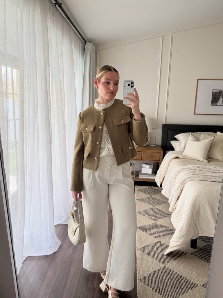 woman in tan cropped jacket and white trousers taking a mirror selfie in bedroom