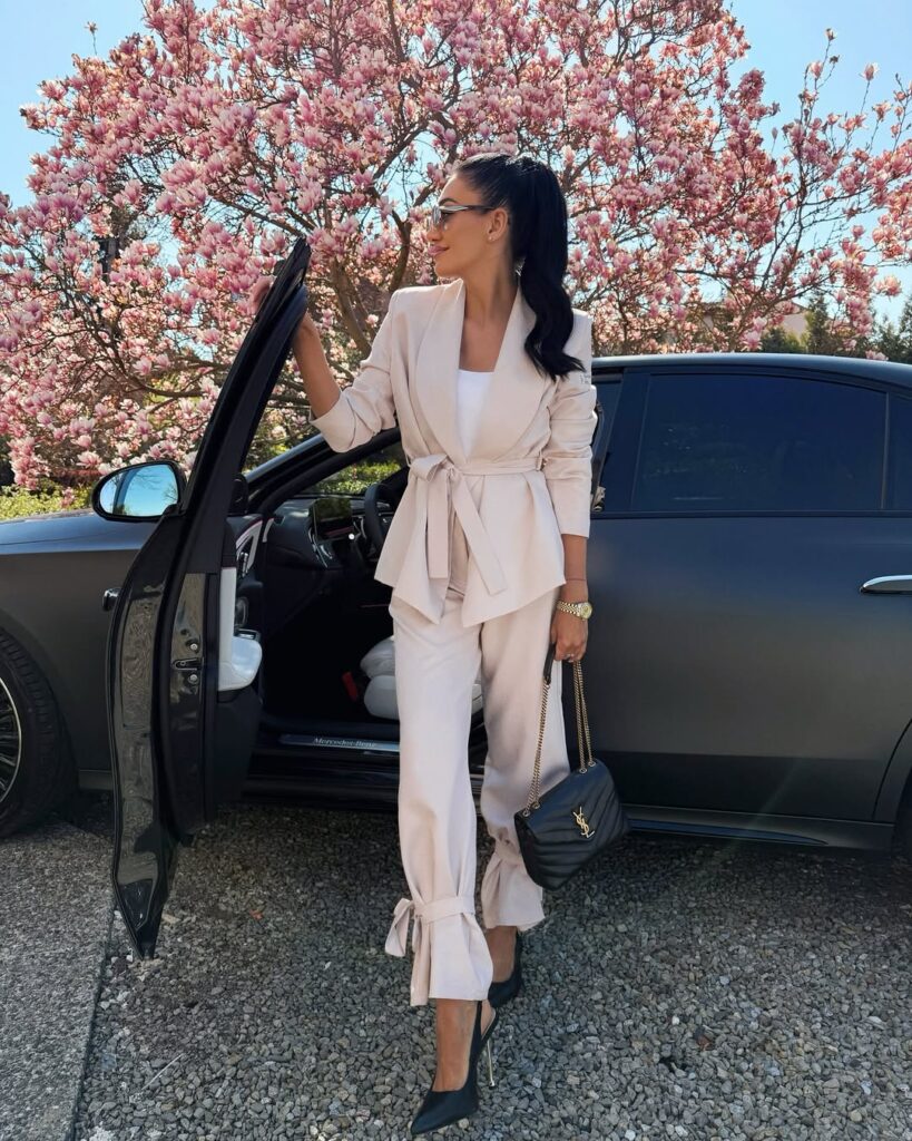 woman in beige belted suit standing by black car near blooming magnolia tree