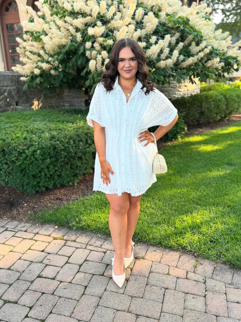woman in white eyelet dress and white heels standing in front of flowering bush